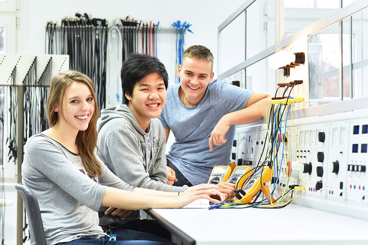 3 young adults in front of a electronic panel smiling at the camera